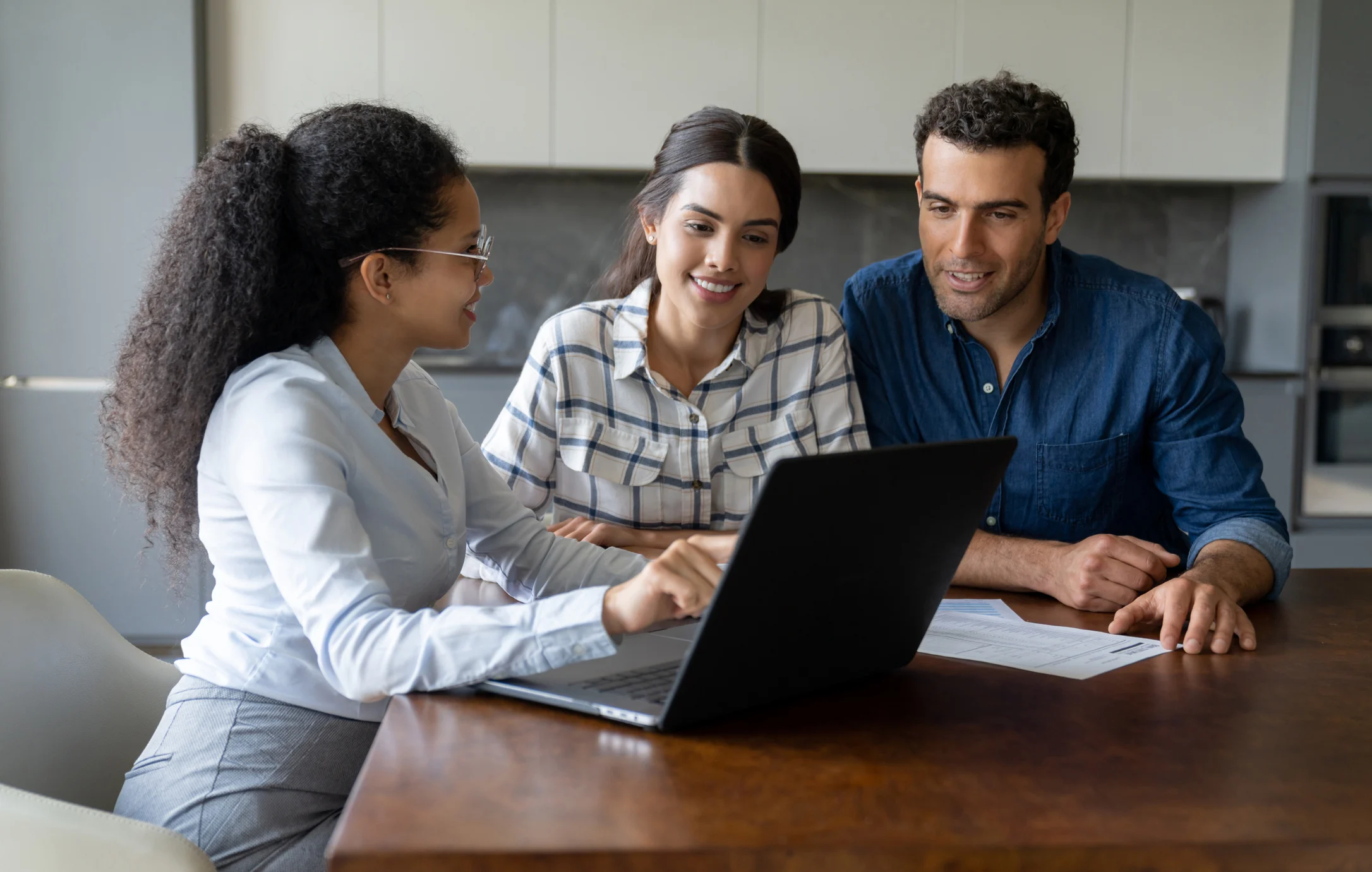 Professional advisor showing digital plans to a couple on a laptop. Bright natural lighting in a modern kitchen with documents on the table.